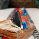 Large multistone inlay ring by Wilbert Grey featuring a rectangular geometric design of orange-red shell, turquoise, and deep blue stones with silver channel accents in a wide polished silver band; side view.