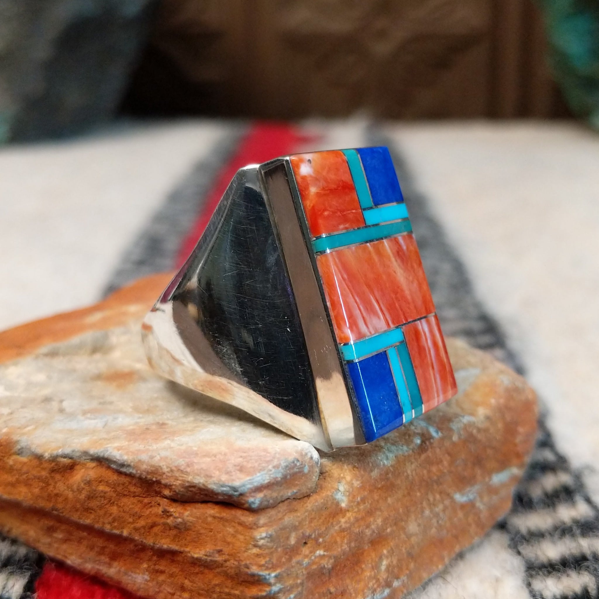 Large multistone inlay ring by Wilbert Grey featuring a rectangular geometric design of orange-red shell, turquoise, and deep blue stones with silver channel accents in a wide polished silver band; side view.