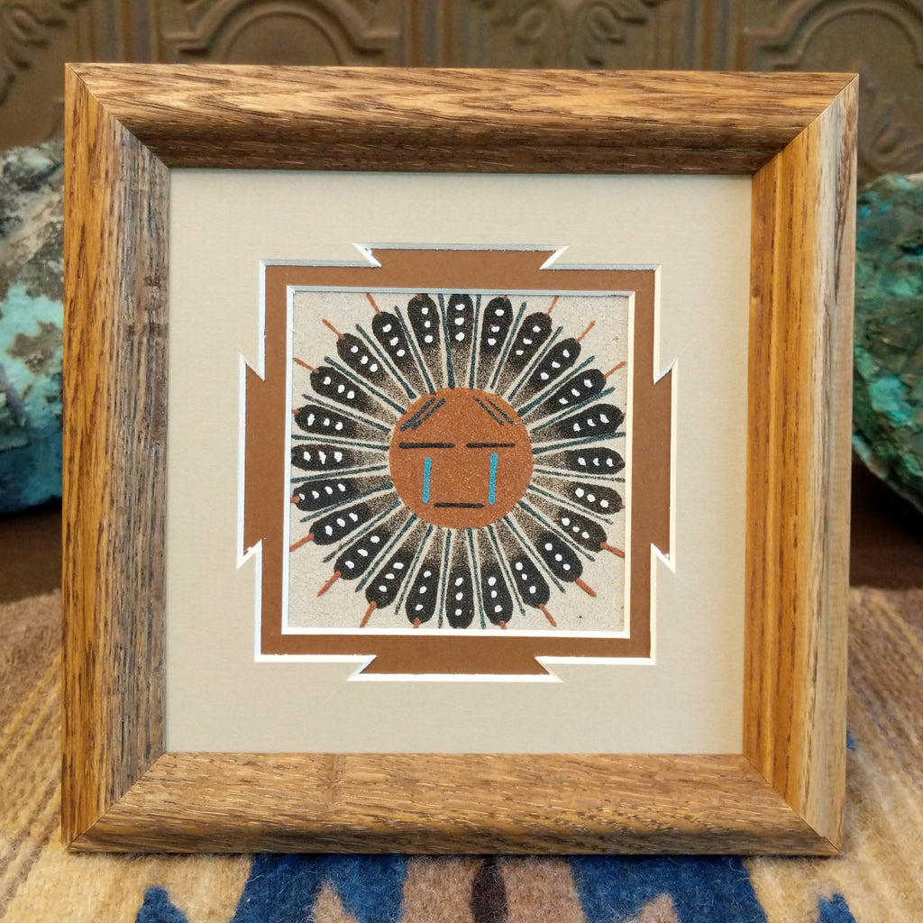 Sand painting of a large Sunface in a wooden frame.
