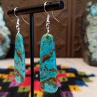 Large turquoise slab earrings by Jameson Pete featuring elongated bright blue stones with bold brown, tan, and dark natural matrix, suspended from silver hooks; side view.