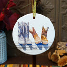Round ceramic Christmas ornament featuring a painted illustration of three cowboy boots in white, tan, and brown, hanging by a gold ribbon against a rustic holiday background.