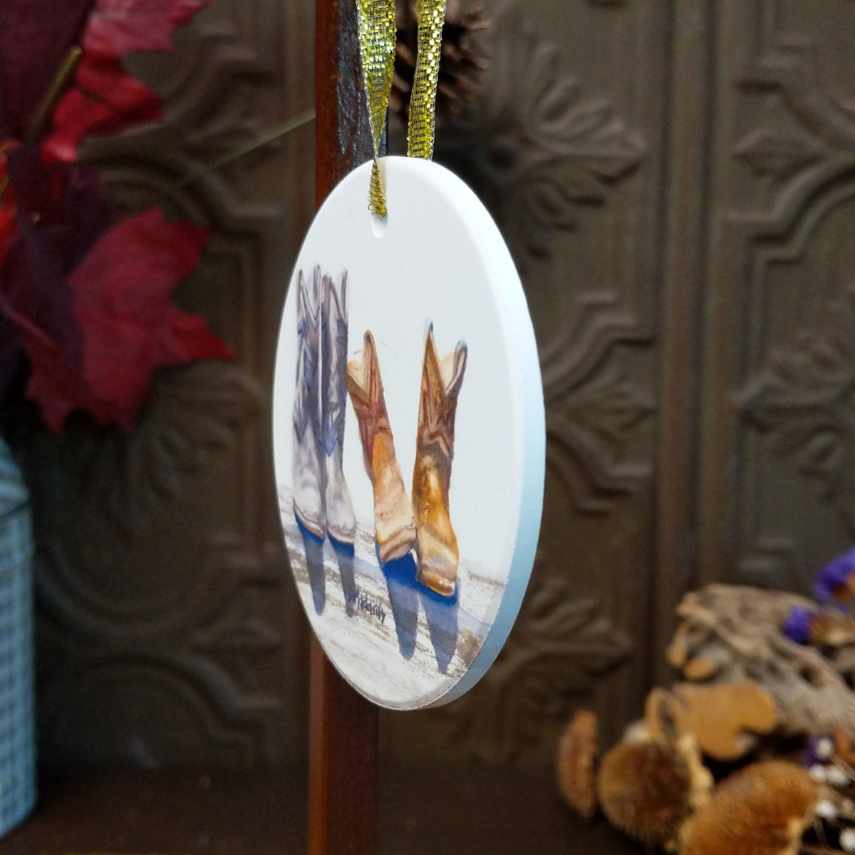 An oval white Christmas ornament featuring a printed illustration of four cowboy boots in different colors, hanging from a gold ribbon against a dark wood background with decorative autumn elements.