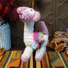 This charming Llama Wool Ornament features a white crocheted body, pastel pink face, ears, hooves, and curled tail, a colorful Southwestern-inspired saddle blanket, black embroidered facial details, and a gold hanging cord.