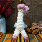 This charming Llama Wool Ornament features a white crocheted body, pastel pink face, ears, hooves, and curled tail, a colorful Southwestern-inspired saddle blanket, black embroidered facial details, and a gold hanging cord.
