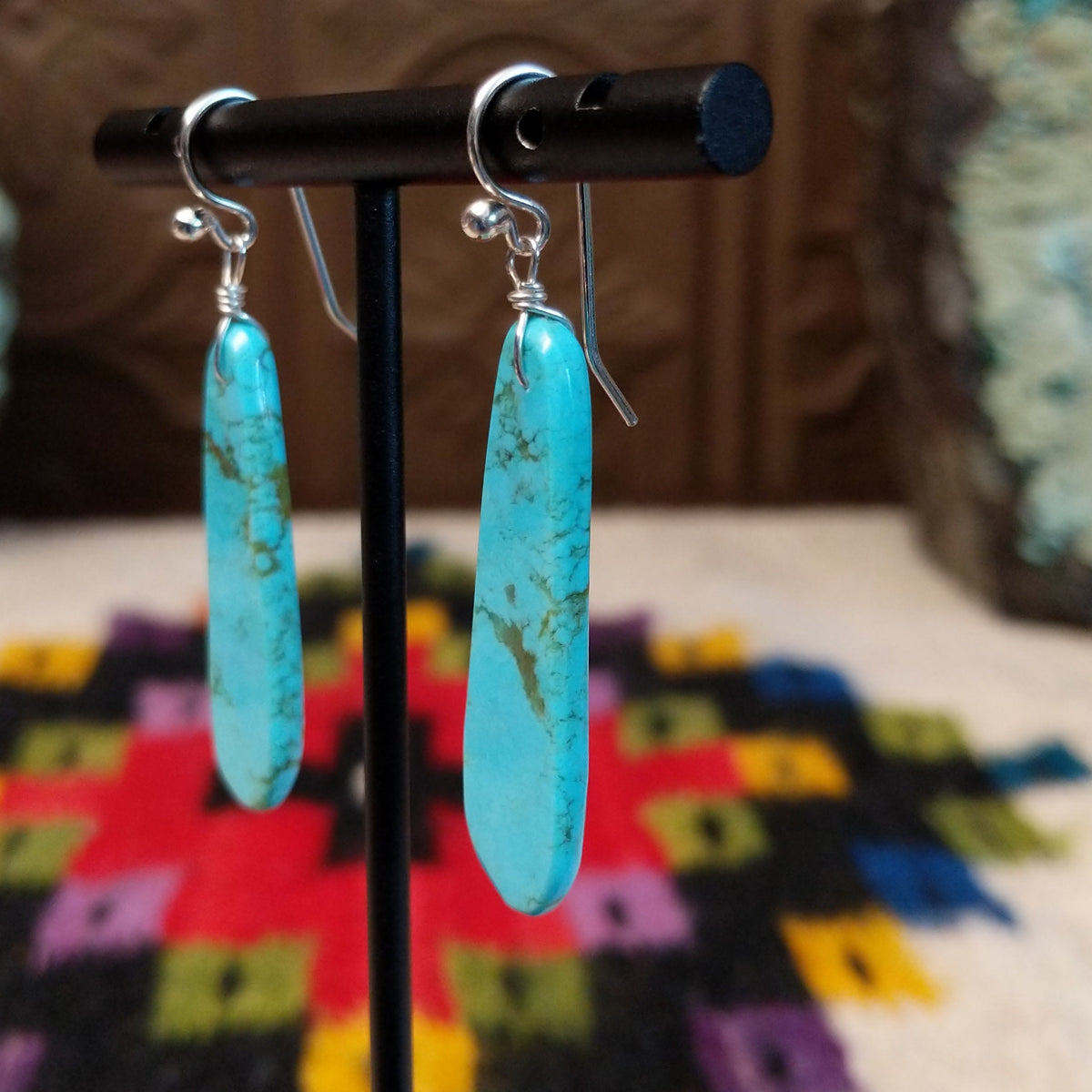 Long Slab Turquoise Earrings by Ronald Chavez featuring elongated bright blue turquoise stones with natural brown and green matrix, suspended from silver hooks; side view.