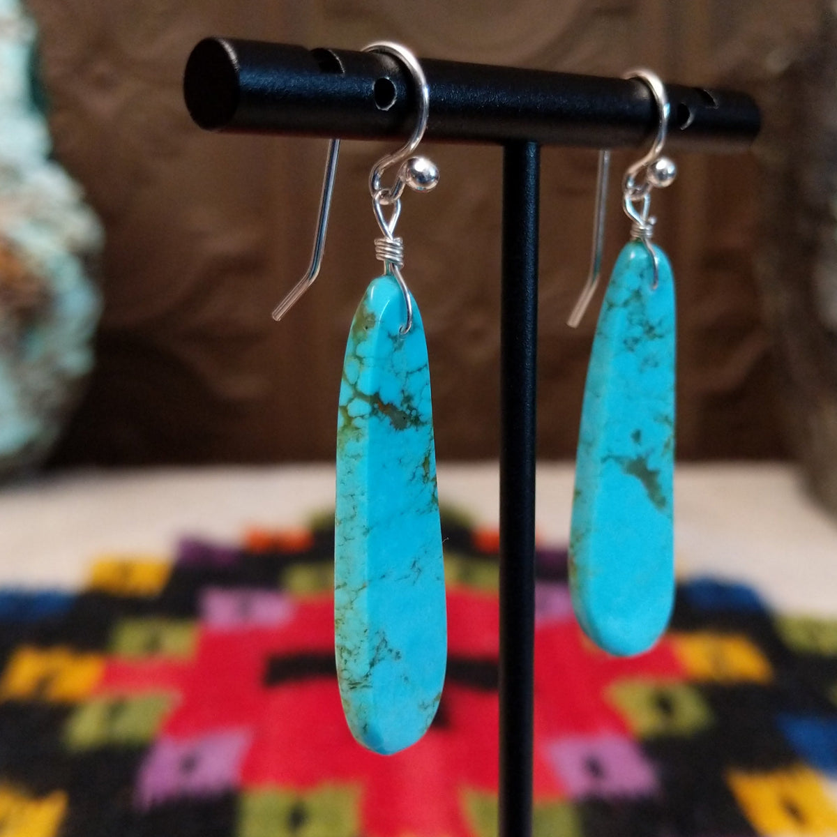 Long Slab Turquoise Earrings by Ronald Chavez featuring elongated bright blue turquoise stones with natural brown and green matrix, suspended from silver hooks; side view.