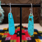 Long Slab Turquoise Earrings by Ronald Chavez featuring elongated bright blue turquoise stones with natural brown and green matrix, suspended from silver hooks; backside view.