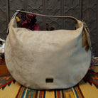 Lulu Handbag by Liz Soto featuring a spacious hobo silhouette, soft suede-like exterior, long shoulder strap, side buckle detailing, top zip closure, and logo plaque.