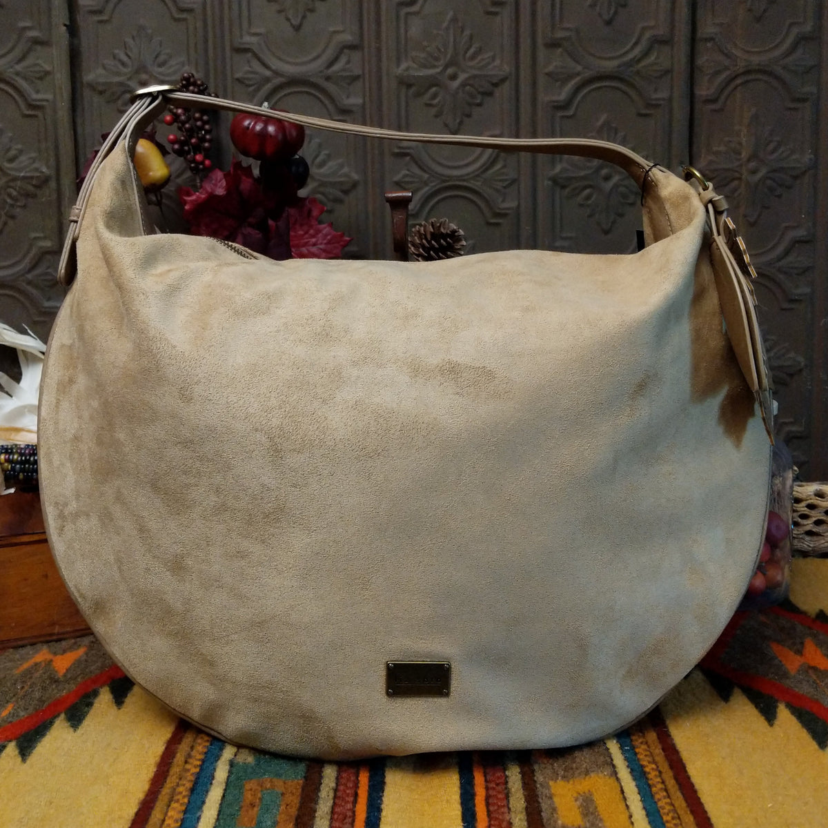 Lulu Handbag by Liz Soto featuring a spacious hobo silhouette, soft suede-like exterior, long shoulder strap, side buckle detailing, top zip closure, and logo plaque.