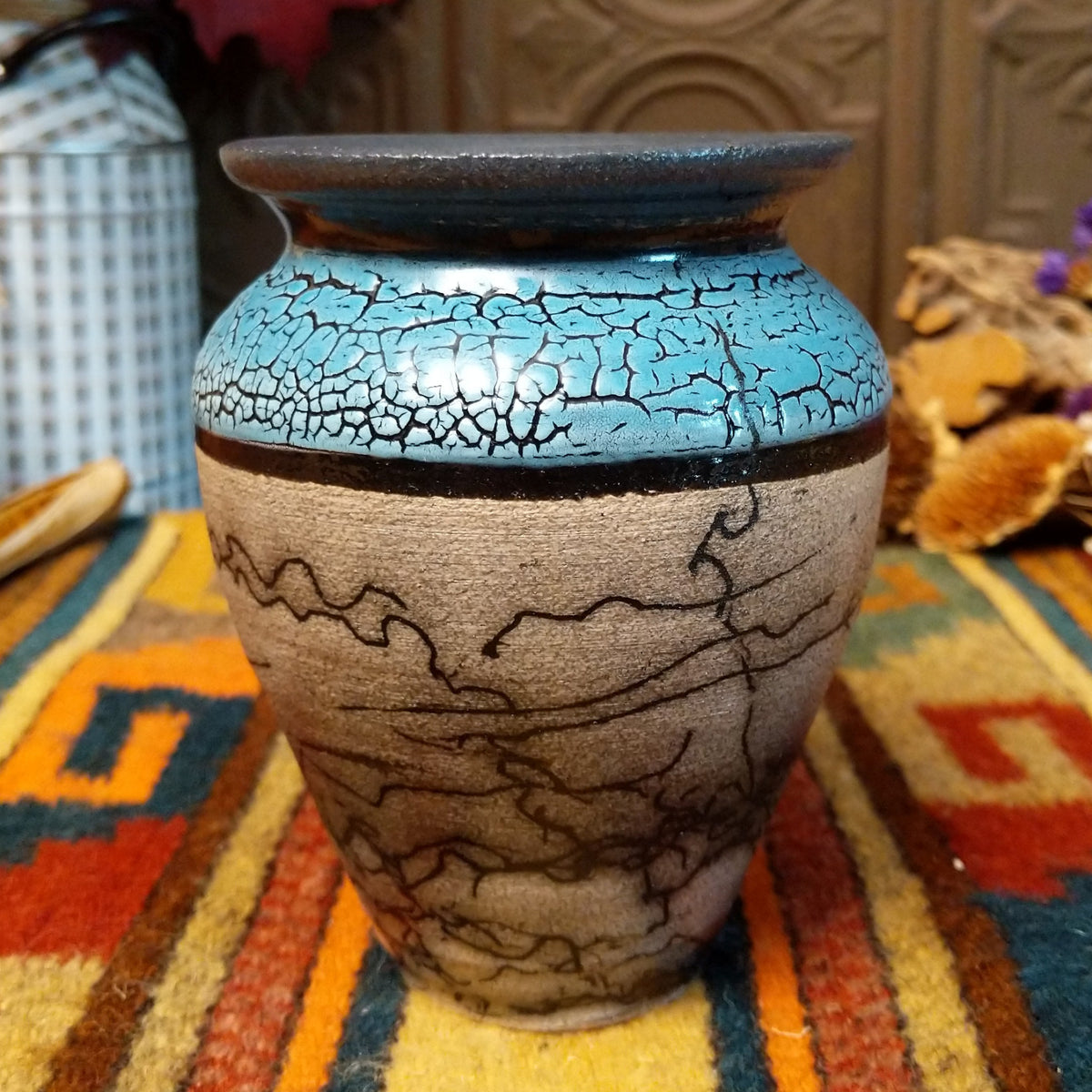 Mini horsehair pottery vase by Roger Calhoun with a flared dark rim, crackled turquoise-blue shoulder, and black horsehair markings concentrated near the base of its cream-colored body.