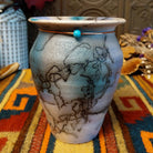 This beautiful Mini Mottled Blue Horsehair Pottery by Roger Calhoun features a small tapered ceramic vase with mottled blue and turquoise tones, black horsehair patterns, a flared rim, and decorative copper tie with a turquoise-colored bead.
