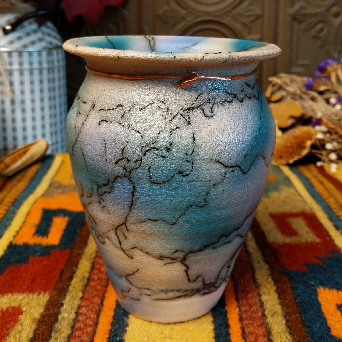 This beautiful Mini Mottled Blue Horsehair Pottery by Roger Calhoun features a small tapered ceramic vase with mottled blue and turquoise tones, black horsehair patterns, a flared rim, and decorative copper tie with a turquoise-colored bead.