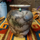 This charming Mini Mottled Green Horsehair Pottery by Roger Calhoun features a rounded vase with a flared black rim, earthy tones, soft green accents, intricate black horsehair patterns, and a copper wire with a green bead.