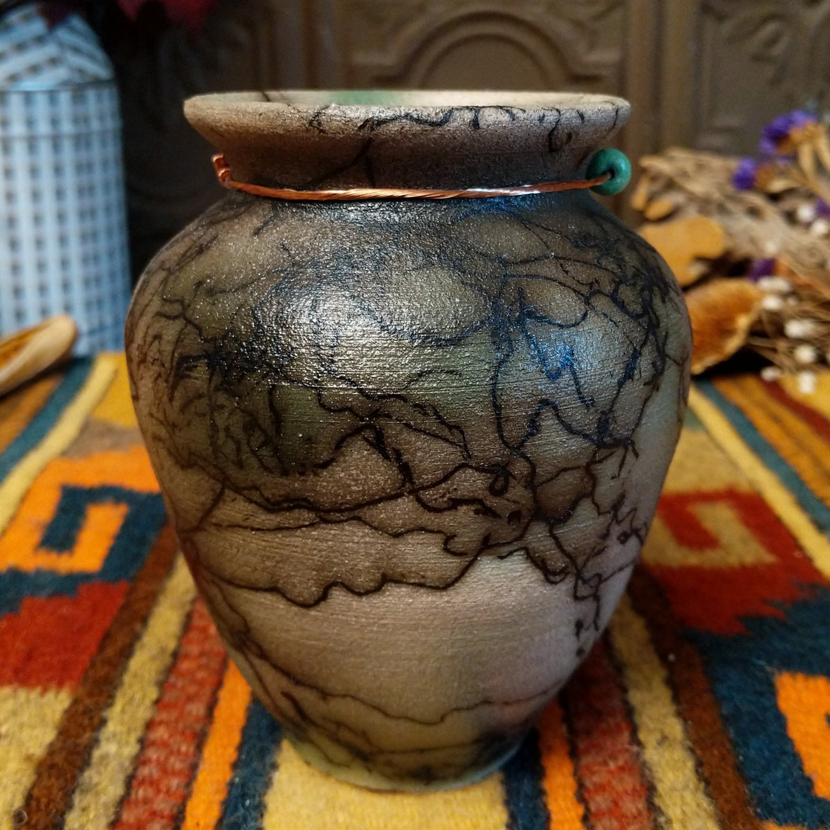 This charming Mini Mottled Green Horsehair Pottery by Roger Calhoun features a rounded vase with a flared black rim, earthy tones, soft green accents, intricate black horsehair patterns, and a copper wire with a green bead.