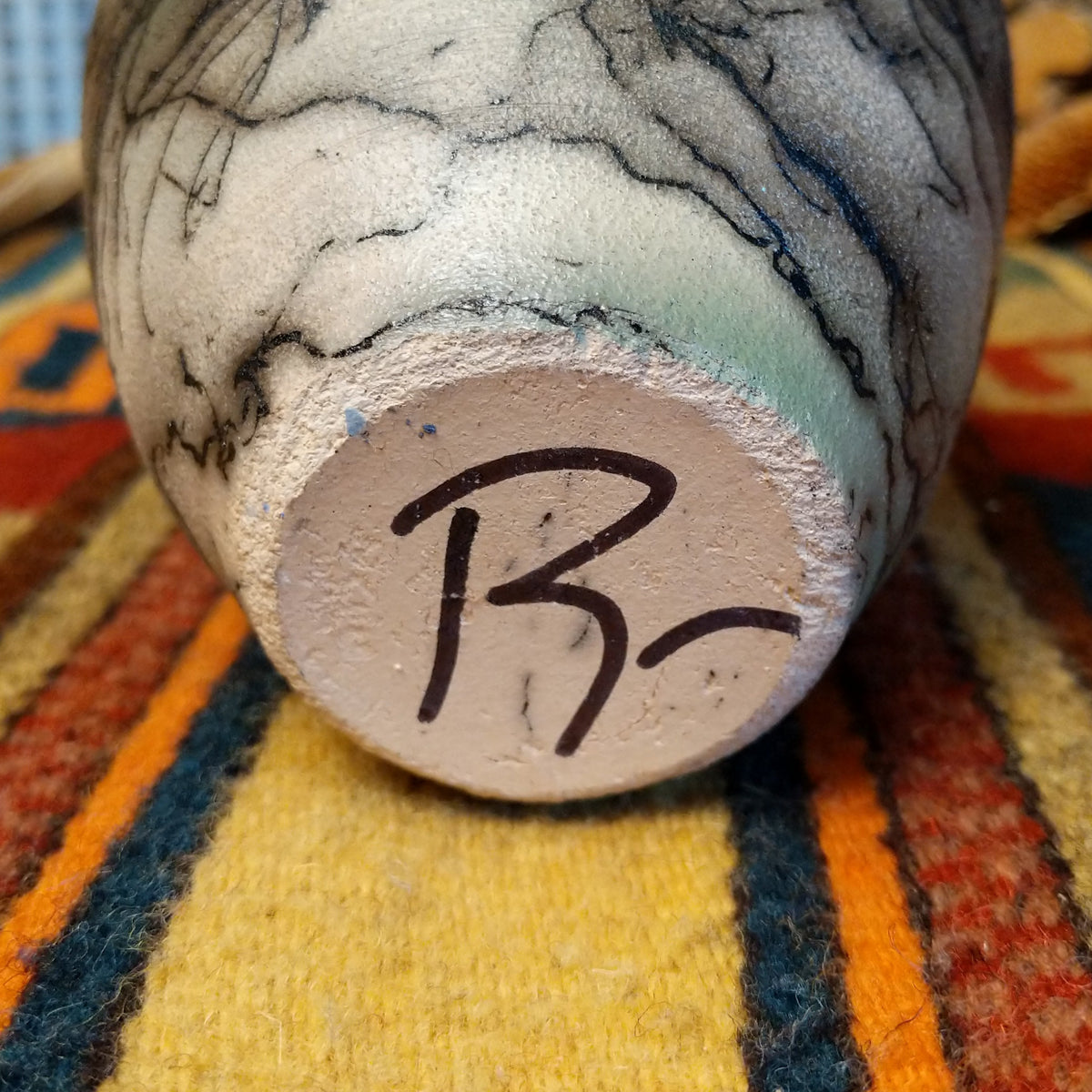 This charming Mini Mottled Green Horsehair Pottery by Roger Calhoun features a rounded vase with a flared black rim, earthy tones, soft green accents, intricate black horsehair patterns, and a copper wire with a green bead; underside and artisan signature view.