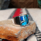 Multistone inlay ring by E.D. featuring geometric turquoise, blue, purple, black, and light-colored stones separated by fine silver channels in a wide polished silver setting; side view.