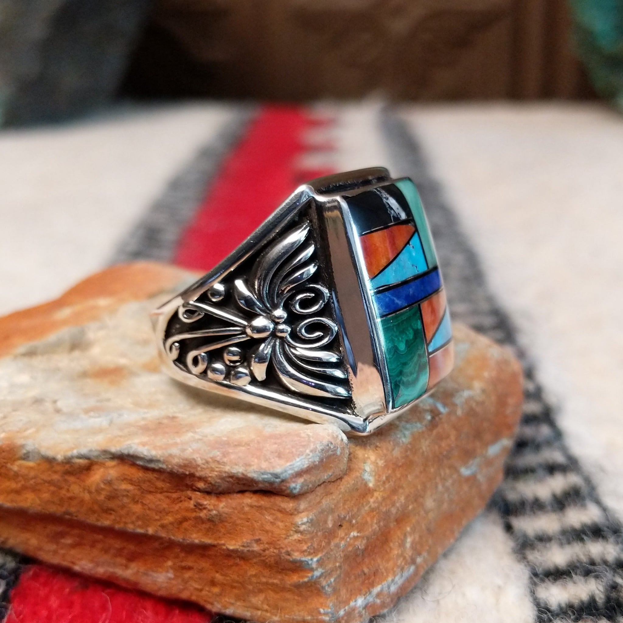 Multistone inlay ring by Terry Wood featuring a rectangular geometric design with turquoise, blue, green, orange, peach, and black stones set in silver with detailed scrollwork along the sides; side view.