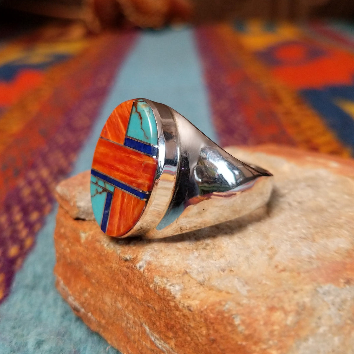 Multistone Inlay Ring by Wilbert Grey featuring an oval geometric design of orange, turquoise, and deep blue stones separated by fine silver channels in a polished silver setting; side view.