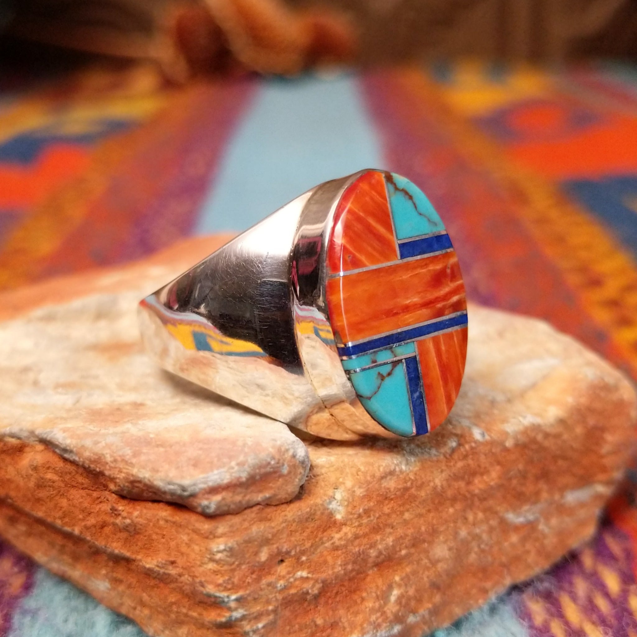Multistone Inlay Ring by Wilbert Grey featuring an oval geometric design of orange, turquoise, and deep blue stones separated by fine silver channels in a polished silver setting; side view.