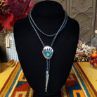 A Sterling Silver bolo tie necklace with a turquoise stone and a paw print pendant, displayed on a black velvet mannequin bust. The necklace features braided blue cords and is set against a rustic background with autumn decorations and a patterned textile.