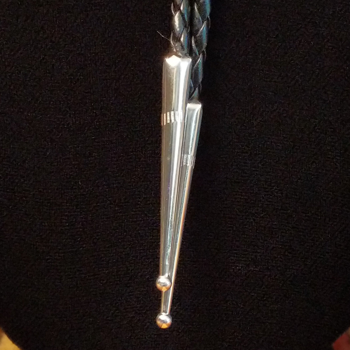 A silver bolo tie necklace with a braided black cord, a turquoise stone, and a polished metal drop, displayed on a black mannequin bust.