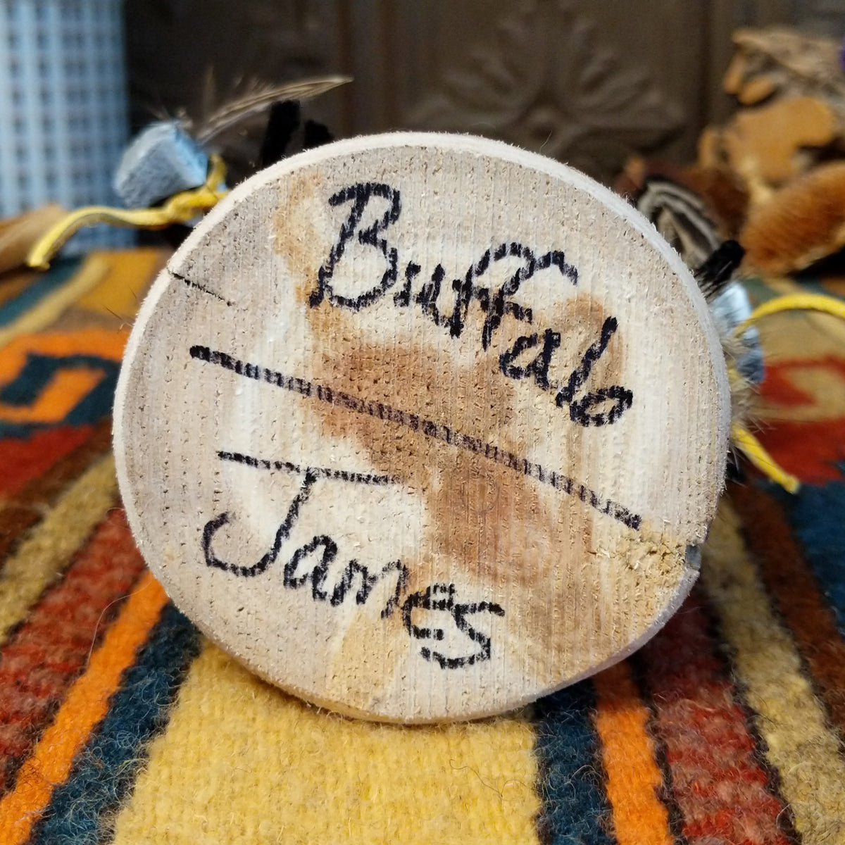 This distinctive Navajo Buffalo Kachina by Debbie James features a hand-painted wooden figure with black buffalo horns, gray fur headdress, black-and-white painted details, natural feathers, leather ties, and a wooden display base; artisan and product name on bottom.
