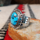 Navajo turquoise ring featuring a large oval bright blue turquoise stone with dark matrix, framed by twisted silver rope and an ornate scalloped silver setting; side view.