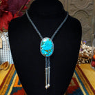 Navajo Kingman Turquoise Bolo Tie featuring a large oval sky-blue turquoise stone with golden-brown matrix, decorative silver bezel, braided black leather cord, and polished silver tips.