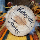 This distinctive Navajo Kokopelli Kachina by Debbie James features a hand-painted wooden figure with a tall feather headdress, black face, white fur accents, natural feathers, leather ties, and a wooden display base; artisan and product name on bottom.