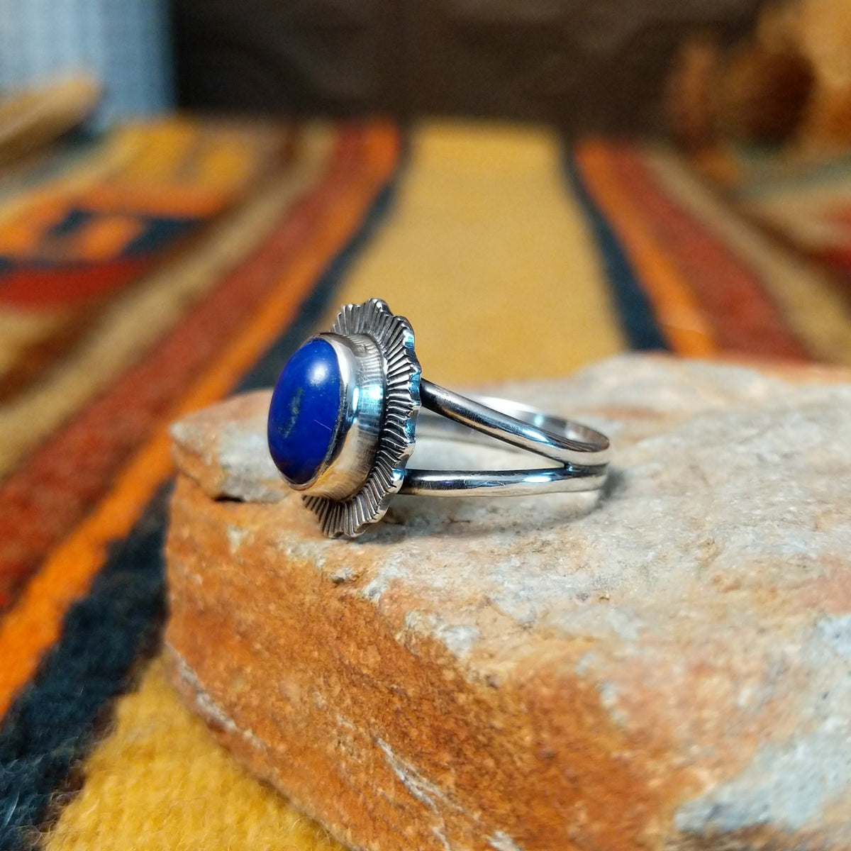 Angled side view of a silver ring featuring a raised oval blue lapis lazuli cabochon, radiating stamped silverwork, and a polished split-shank band.