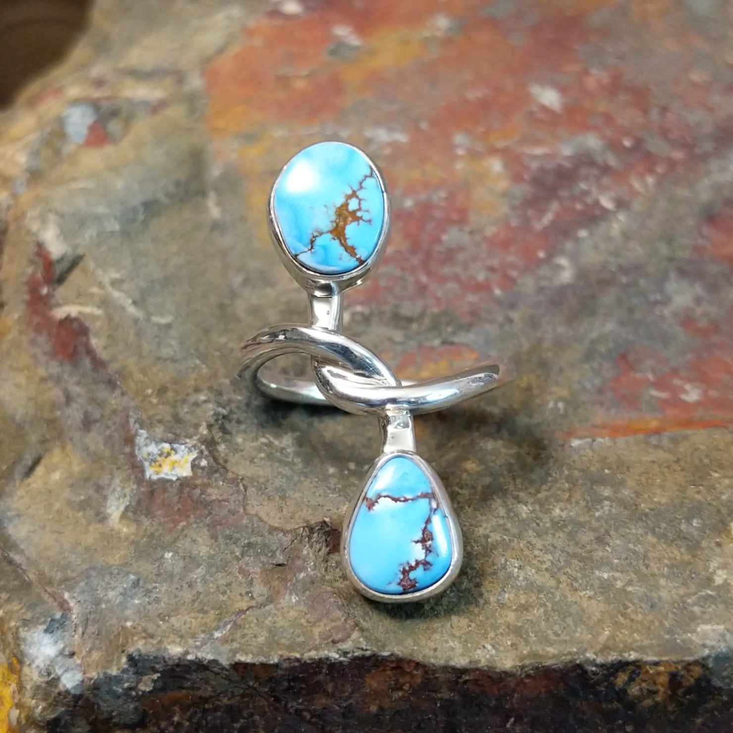 Silver ring with entwined band holding an oval turquoise stone and a teardrop turquoise stone in smooth bezels. Matrix veining visible in both stones.