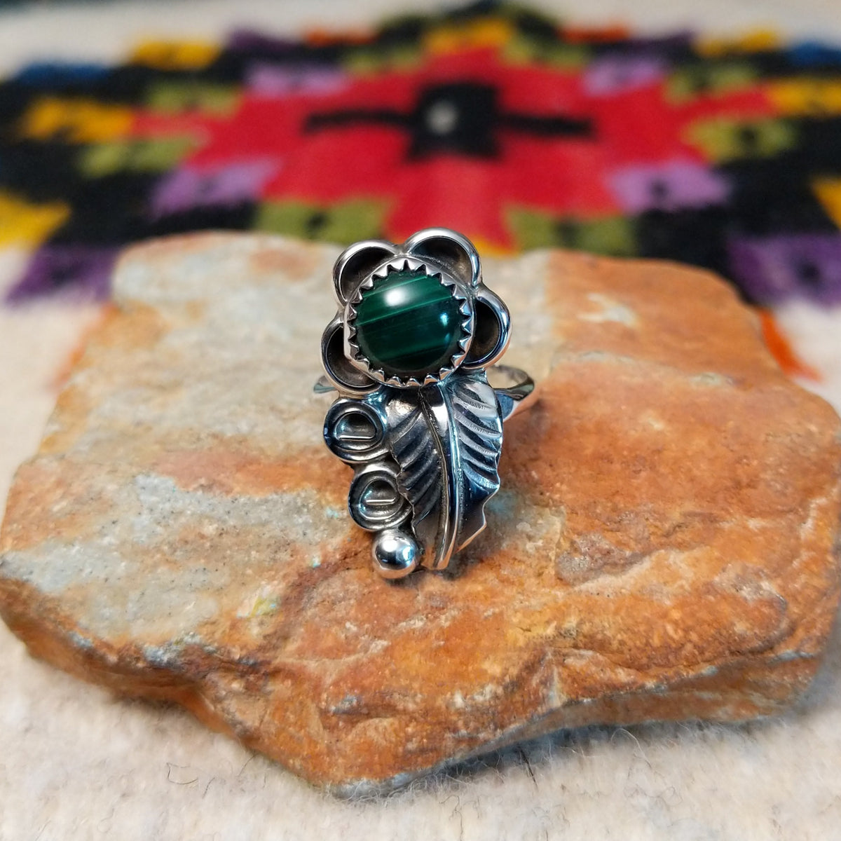 This Navajo Malachite Feather Ring by R.M. features a round green malachite stone in sterling silver surrounded by floral accents, detailed feathers, silver scrolls, and polished beadwork.