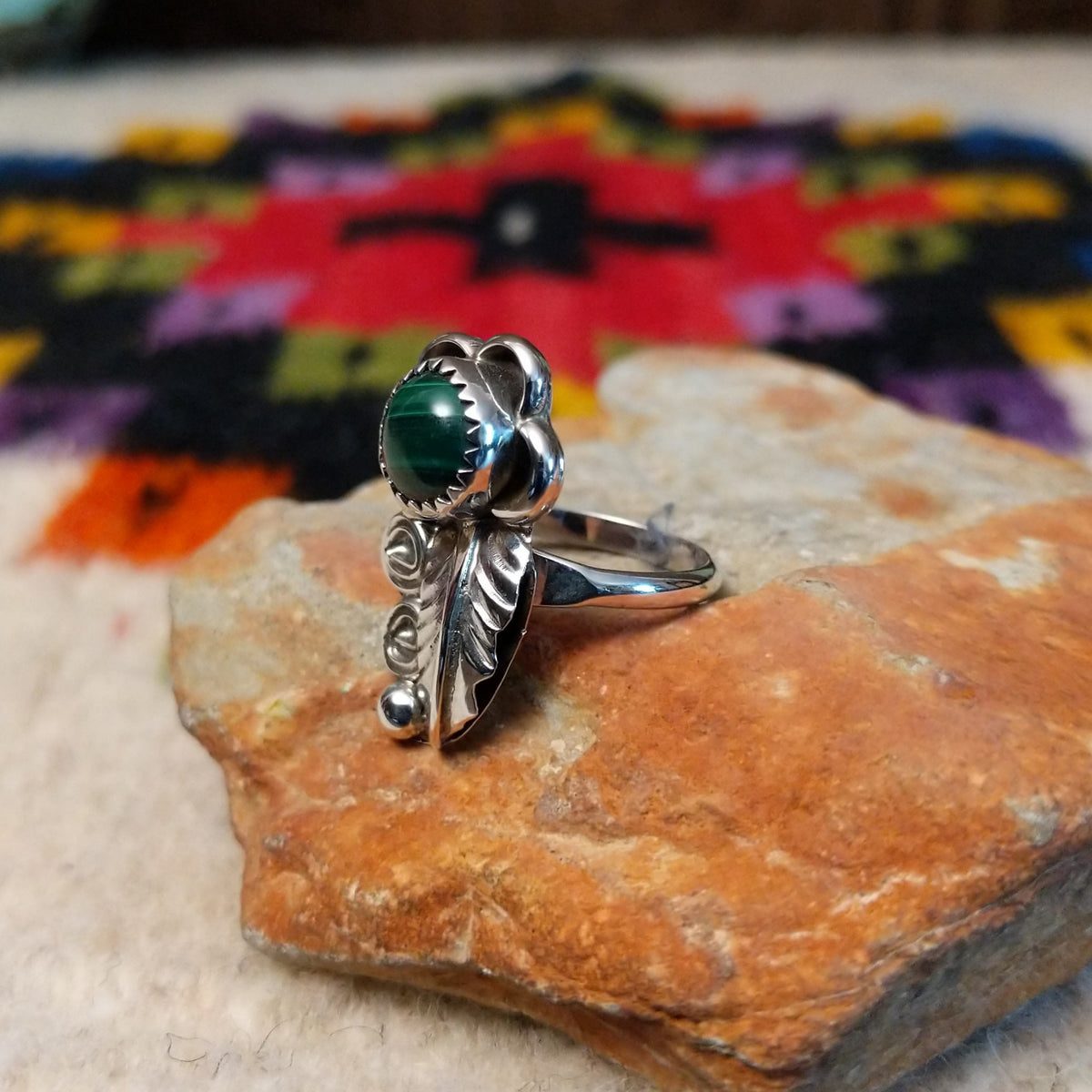 This Navajo Malachite Feather Ring by R.M. features a round green malachite stone in sterling silver surrounded by floral accents, detailed feathers, silver scrolls, and polished beadwork; side view.
