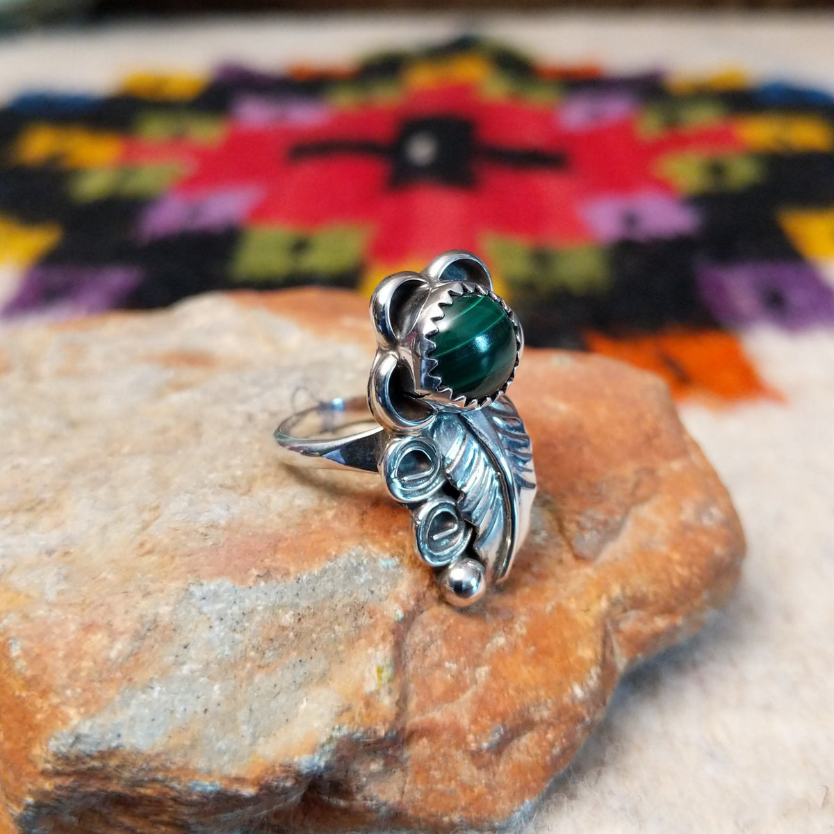 This Navajo Malachite Feather Ring by R.M. features a round green malachite stone in sterling silver surrounded by floral accents, detailed feathers, silver scrolls, and polished beadwork; side view.