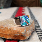 Navajo Multistone Inlay Ring by A.M.P. featuring a square geometric mosaic of turquoise, blue, purple, orange, red, and iridescent inlay set in a wide polished silver band; side view.