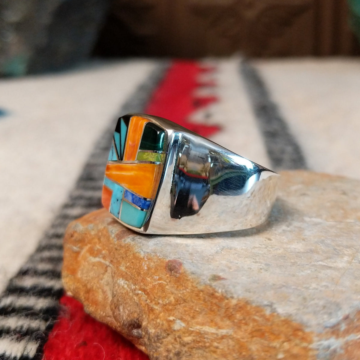 Navajo multistone inlay ring by O.J. featuring a rectangular geometric design with orange shell, turquoise, blue, green, and black inlay set in a polished silver band; side view.