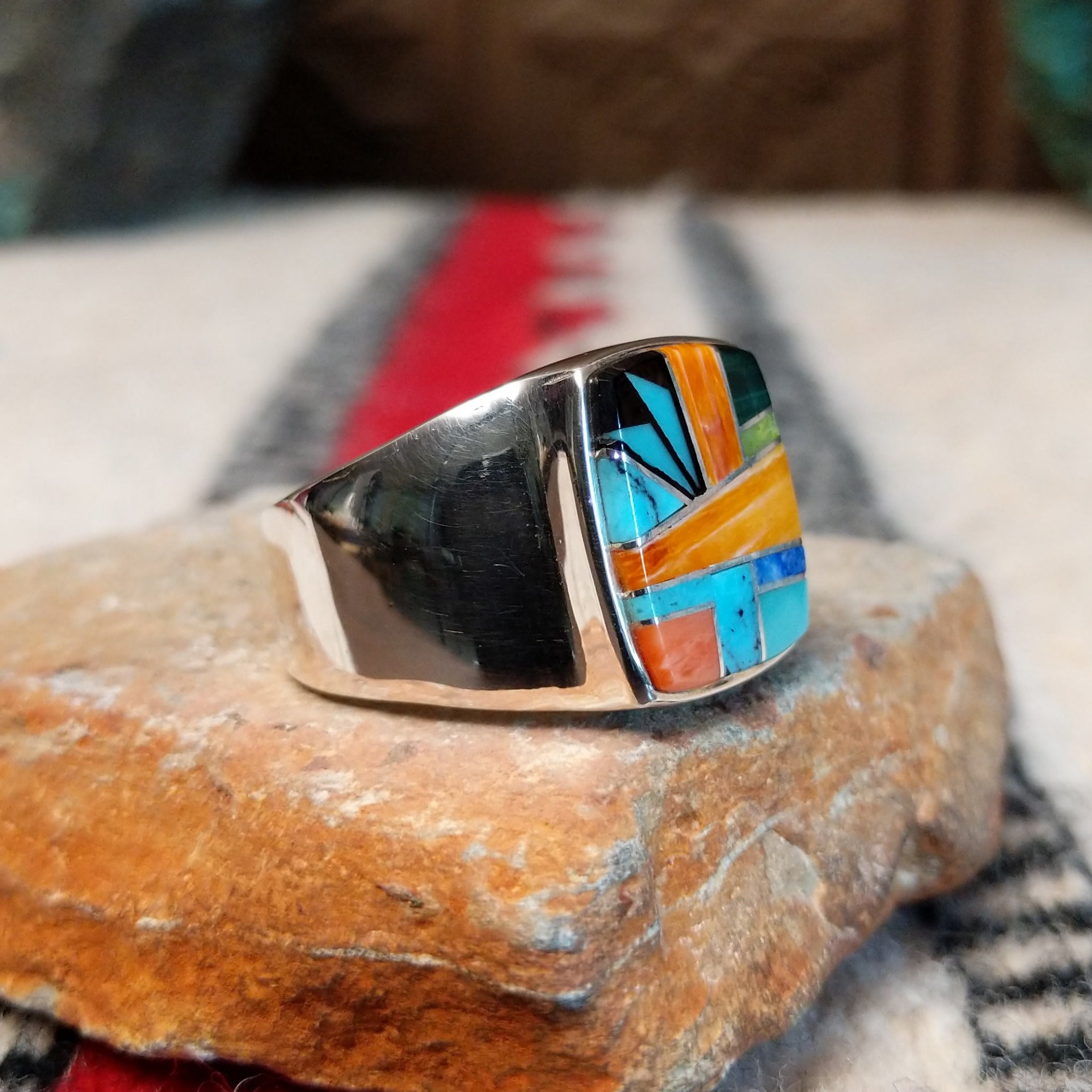 Navajo multistone inlay ring by O.J. featuring a rectangular geometric design with orange shell, turquoise, blue, green, and black inlay set in a polished silver band; side view.