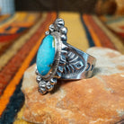 A silver ring with a large oval turquoise stone, displayed on a small rock over a patterned textile.