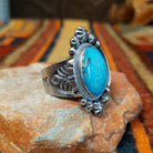 This elegant Navajo Ornate Sonoran Gold Turquoise Ring features a large oval blue turquoise stone surrounded by a silver rope border, polished silver beads, and decorative stamped silver blossoms; side view.