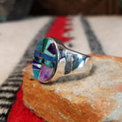 Navajo oval multistone inlay ring by E.D. featuring turquoise, blue, green, and purple stones arranged in a geometric mosaic with silver channel accents and a polished silver band; side view.