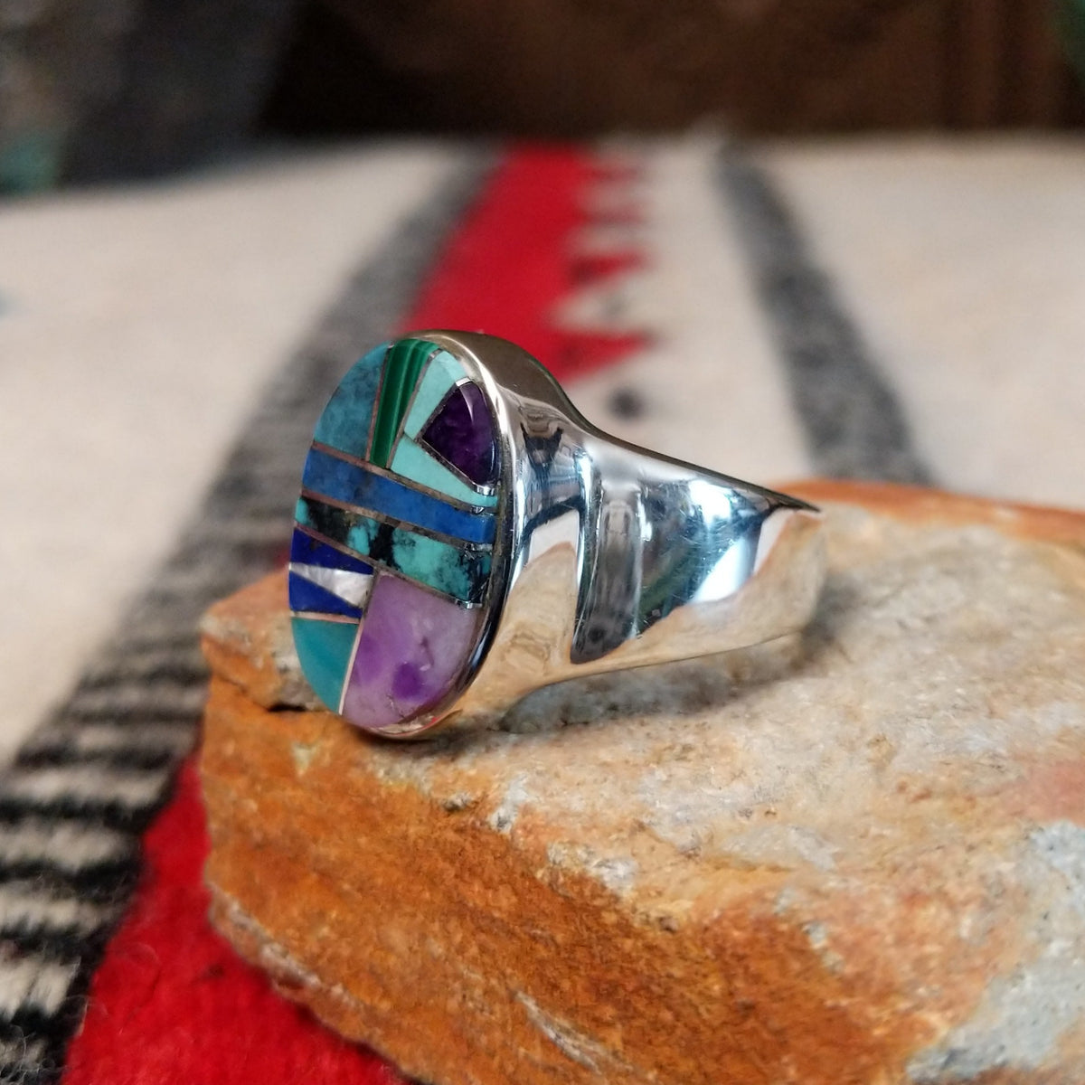 Navajo oval multistone inlay ring by E.D. featuring turquoise, blue, green, and purple stones arranged in a geometric mosaic with silver channel accents and a polished silver band; side view.