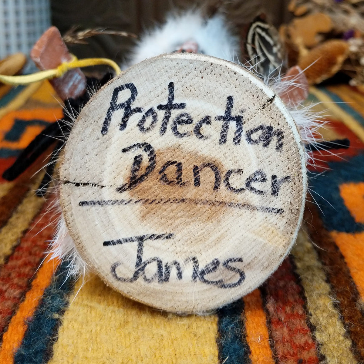 This distinctive Navajo Protection Dancer Kachina by Debbie James features a hand-painted wooden figure with a white feather headdress, natural feathers, leather ties, fur accents, colorful fabric details, and a wooden display base; name and artisan name on bottom.