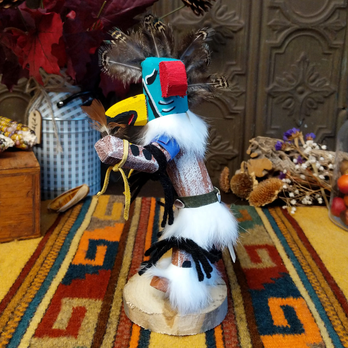 This colorful Navajo Quail Kachina by Debbie James features a turquoise hand-painted bird mask with red accents, a yellow beak, natural feather crest, white fur, leather ties, and a wooden display base; side view.