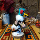 This colorful Navajo Quail Kachina by Debbie James features a turquoise hand-painted bird mask with red accents, a yellow beak, natural feather crest, white fur, leather ties, and a wooden display base; side view.