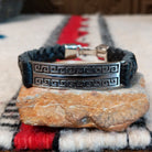 Navajo bracelet featuring braided black leather and a rectangular silver centerpiece with two rows of repeating geometric scroll patterns and darkened recessed details.