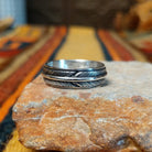 Silver feather-patterned band ring featuring diagonal engraved lines, darkened recesses, and a raised polished silver ridge around the center.