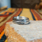 Silver feather-patterned band ring featuring diagonal engraved lines, darkened recesses, and a raised polished silver ridge around the center.