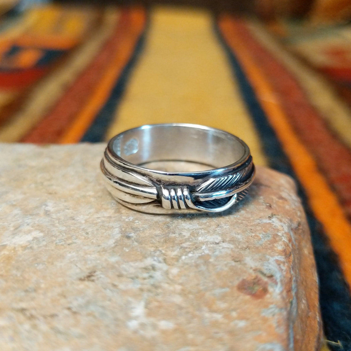 Silver feather-patterned band ring featuring diagonal engraved lines, darkened recesses, and a raised polished silver ridge around the center.