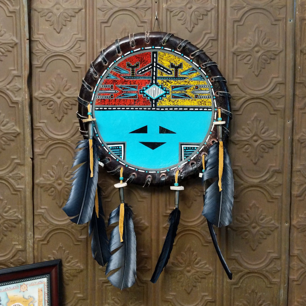Red and yellow paint forms in decorative traditional designs on the base of the painted turquoise sunface with leather ties and dangling black feathers.