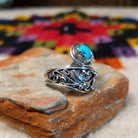 This eye-catching Navajo Turquoise & Coral Feather Ring features turquoise and red coral stones in sawtooth silver bezels with detailed feather, scroll, and silver bead accents; side view.